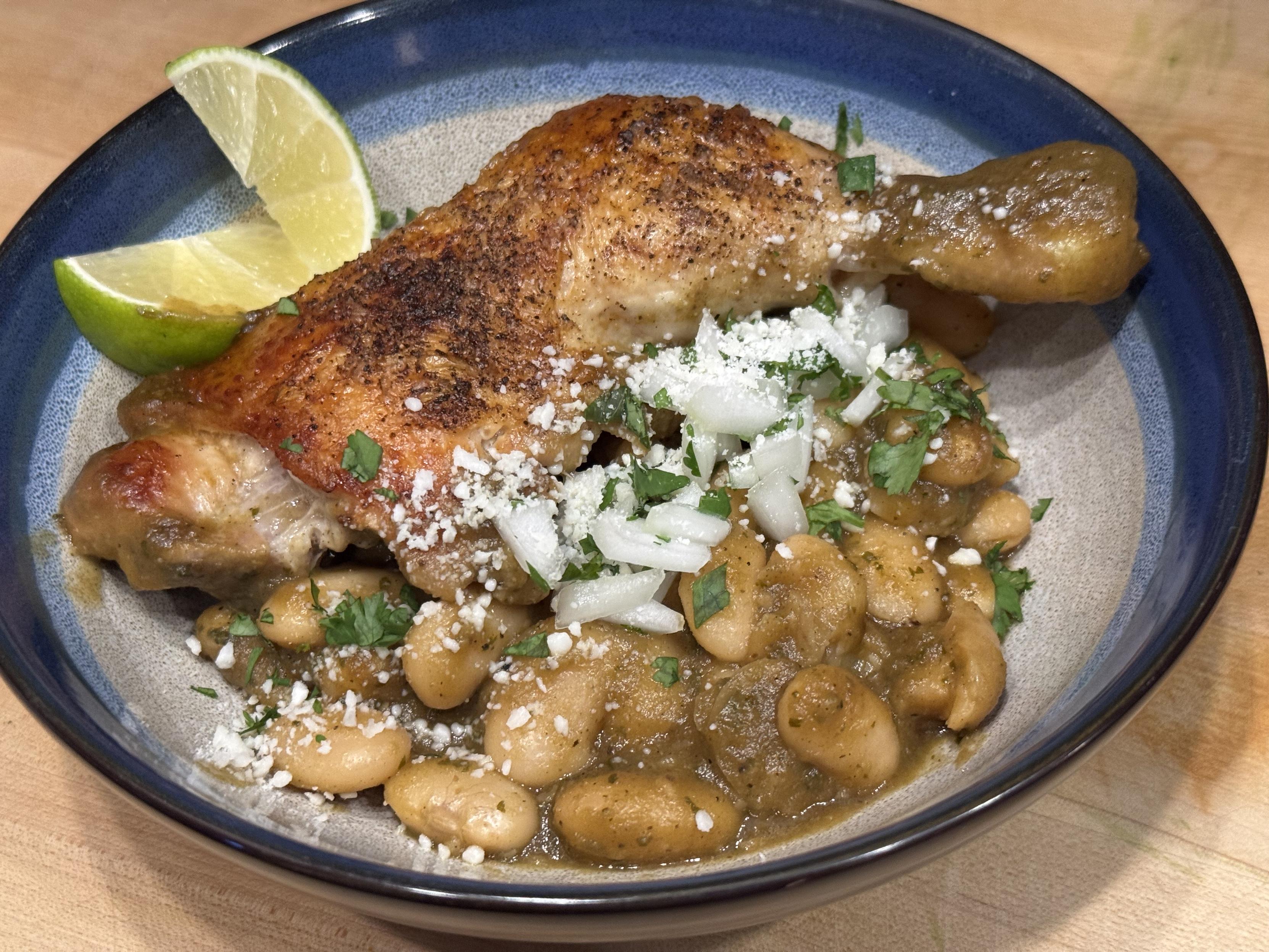 A bowl with a crispy braised chicken leg on top of white beans in a green Chile sauce. Granished with lime wedges, diced onions, and cotija cheese.